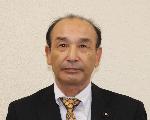 白い背景に、黒いスーツと黄銅色のネクタイを着用した小松栄喜さんの顔写真