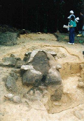 掘り起こされて地中から石の建造物が露出している前方後円墳と、見学者が奥に立っている写真