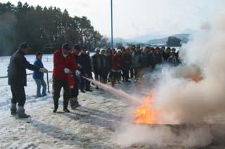 雪が積もっている屋外で、赤い防寒着を着て消火活動を行う男性と、並んで見学する人たちの写真